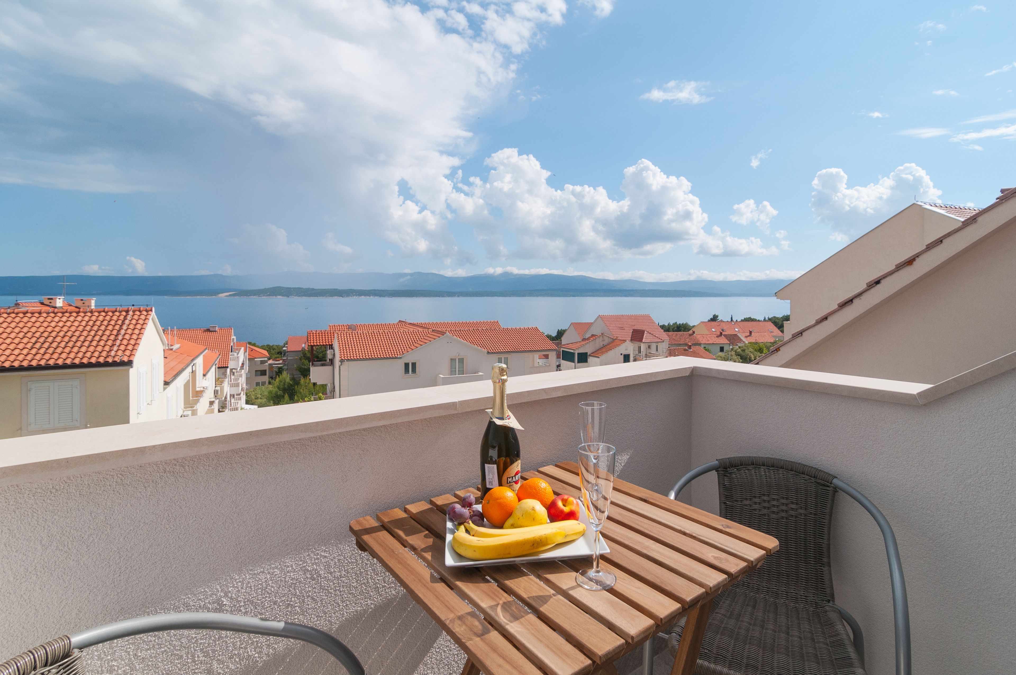 A balcony with a table set for two, featuring a bottle of champagne, a glass, and a fruit plate with bananas, grapes, and oranges.