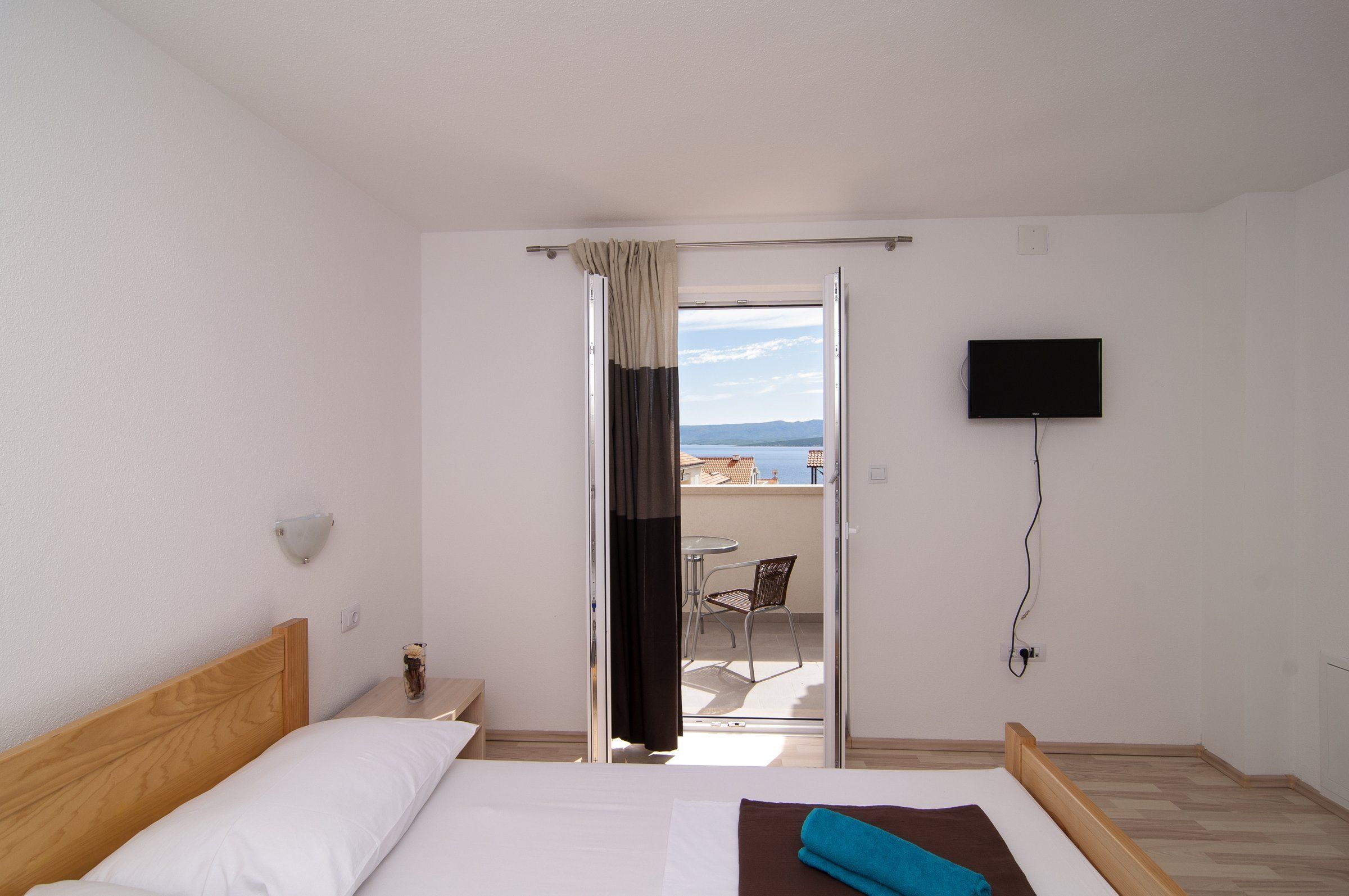 A hotel room with a bed, a TV mounted on the wall, a window with curtains, and a view of the outside balcony.