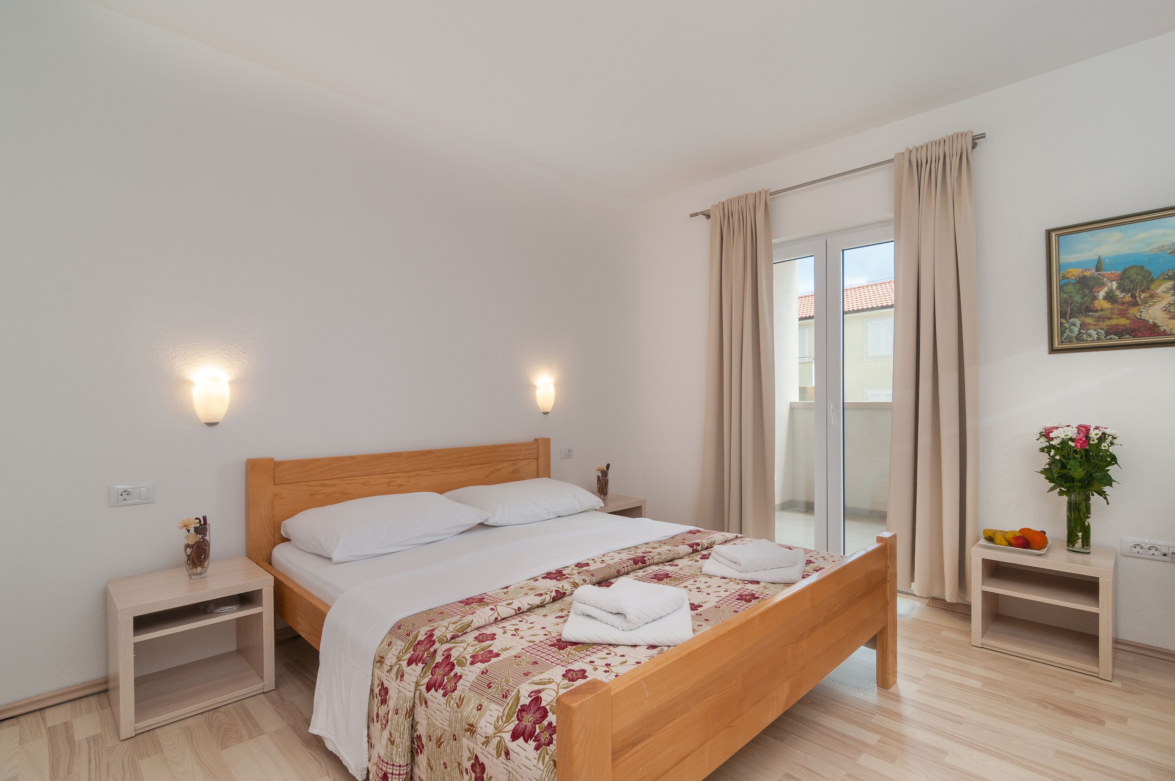A bedroom with a wooden bed, white linens, a nightstand with a vase, and a painting on the wall.