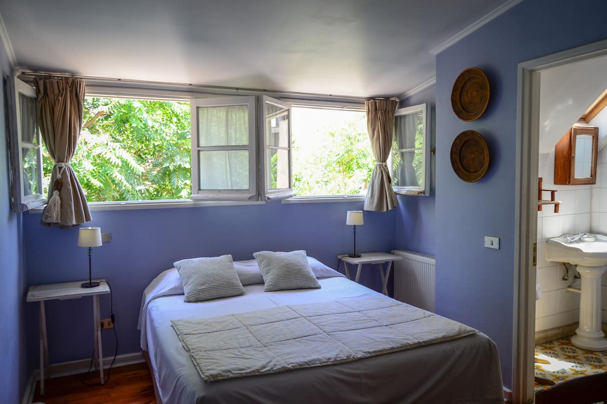 Un dormitorio con cama, dos mesitas de noche, una ventana con cortinas y un baño con lavamanos y espejo.