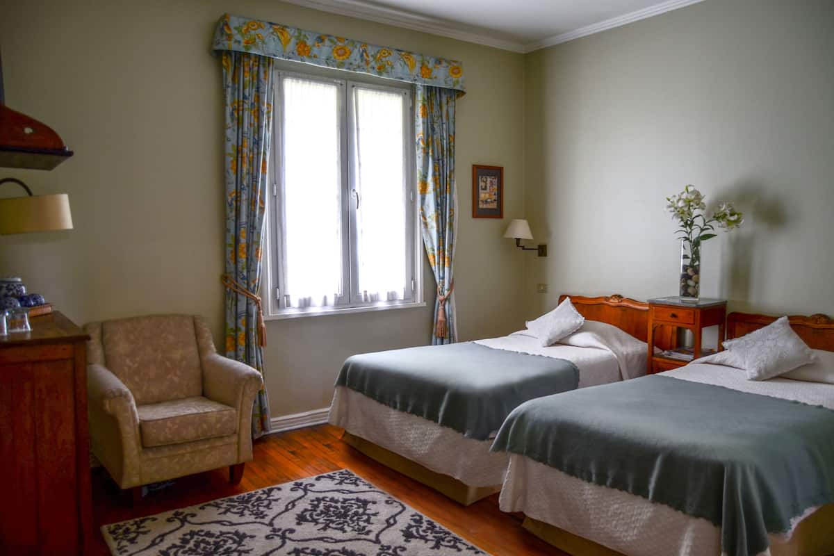 Un dormitorio con dos camas, una silla, una mesita de noche, una lámpara y un ventanal con cortinas.
