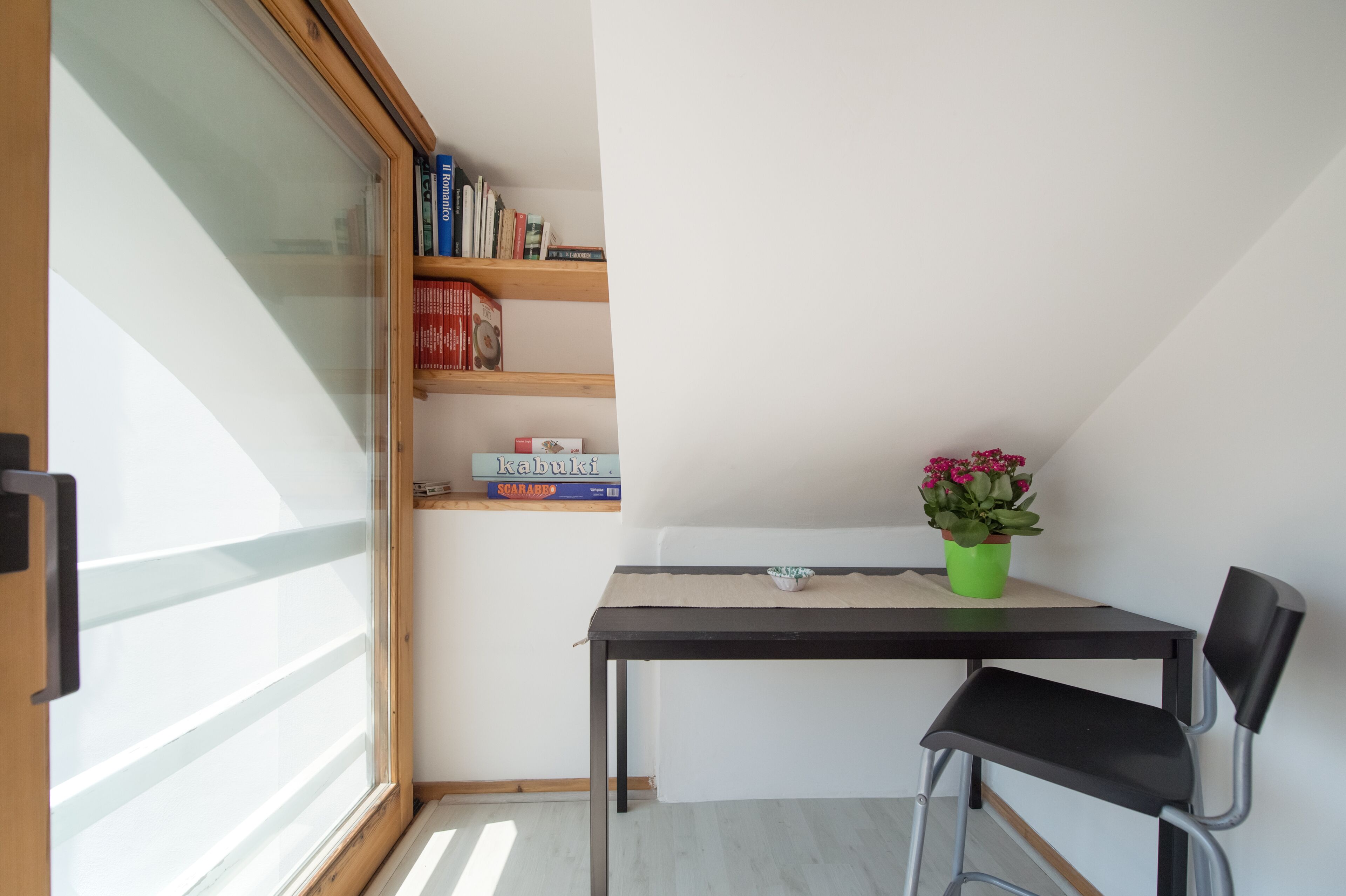 Ein kleines Zimmer mit Fenster, einem Bücherregal, einem Tisch mit Stuhl und einem Blumentopf.