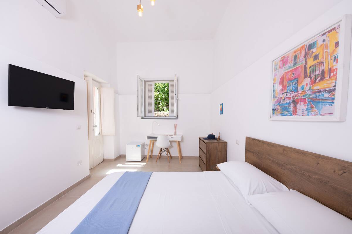 Un dormitorio con cama, escritorio, silla, armario, televisor y ventana con vista a follaje.