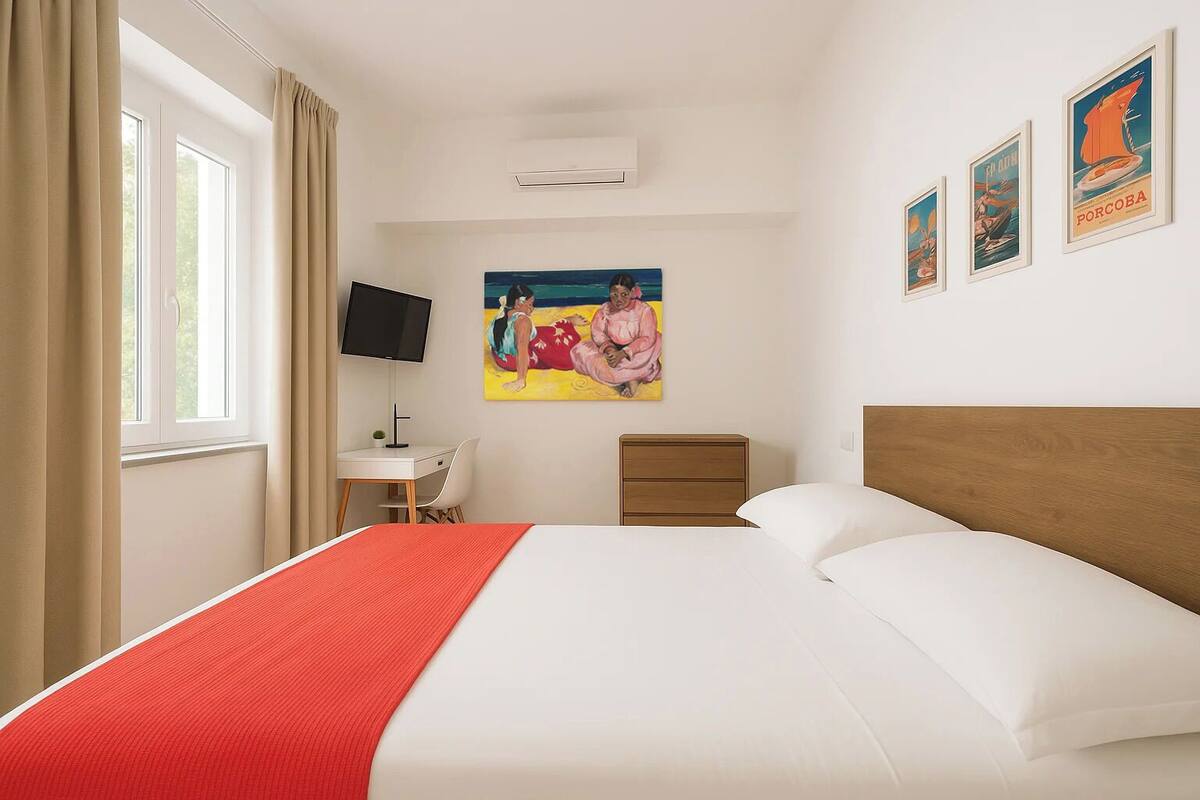 Habitación de hotel con cama, escritorio, silla, televisor, mesita de noche y un cuadro en la pared.