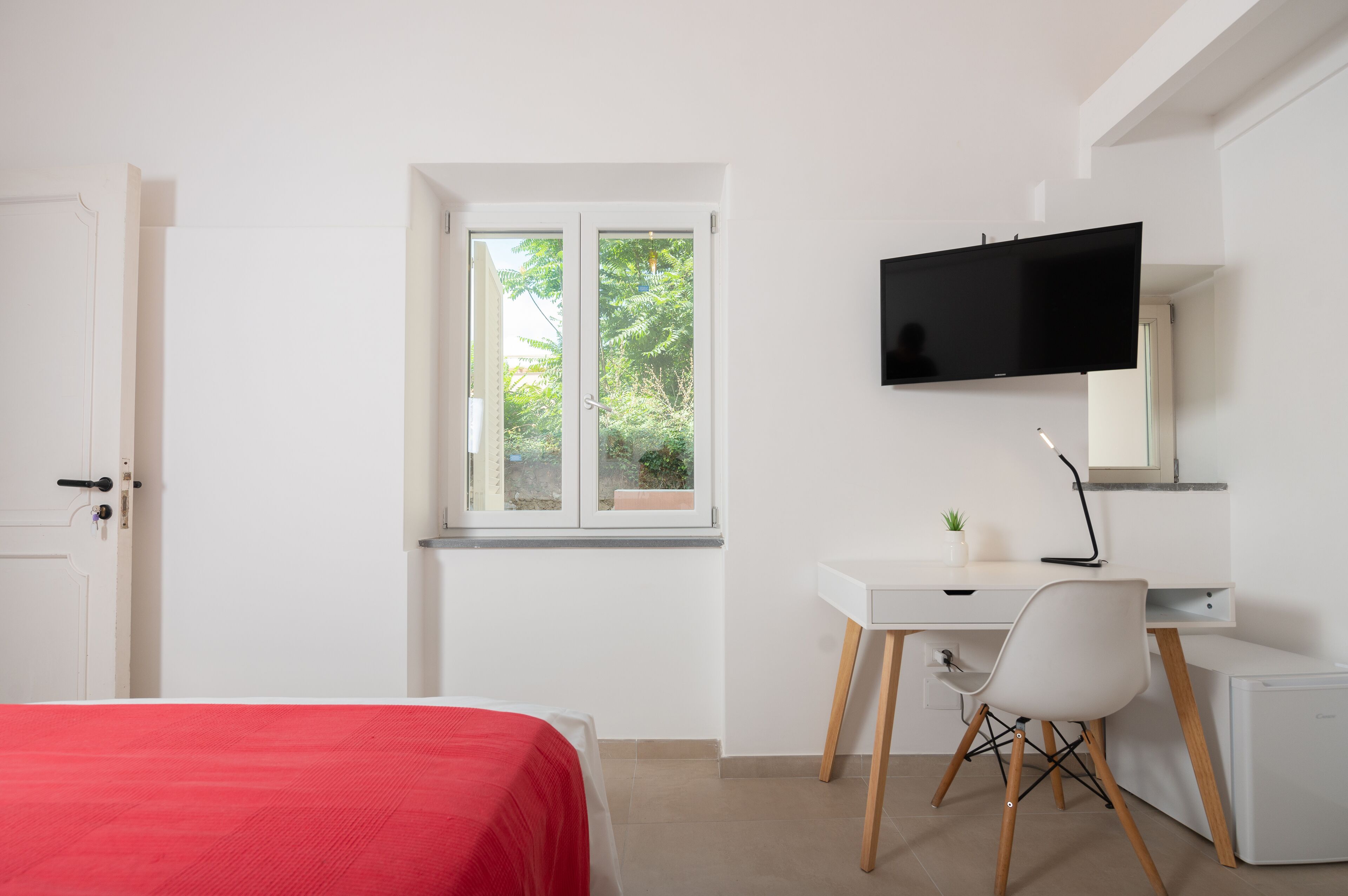 A bedroom with a bed, a desk, a chair, a television, and a window with a view of greenery.