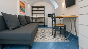Un espace de vie compact comprenant un canapé gris, une table à manger en bois, une chaise, un téléviseur fixé au mur et un coin cuisine avec des étagères et un réfrigérateur.