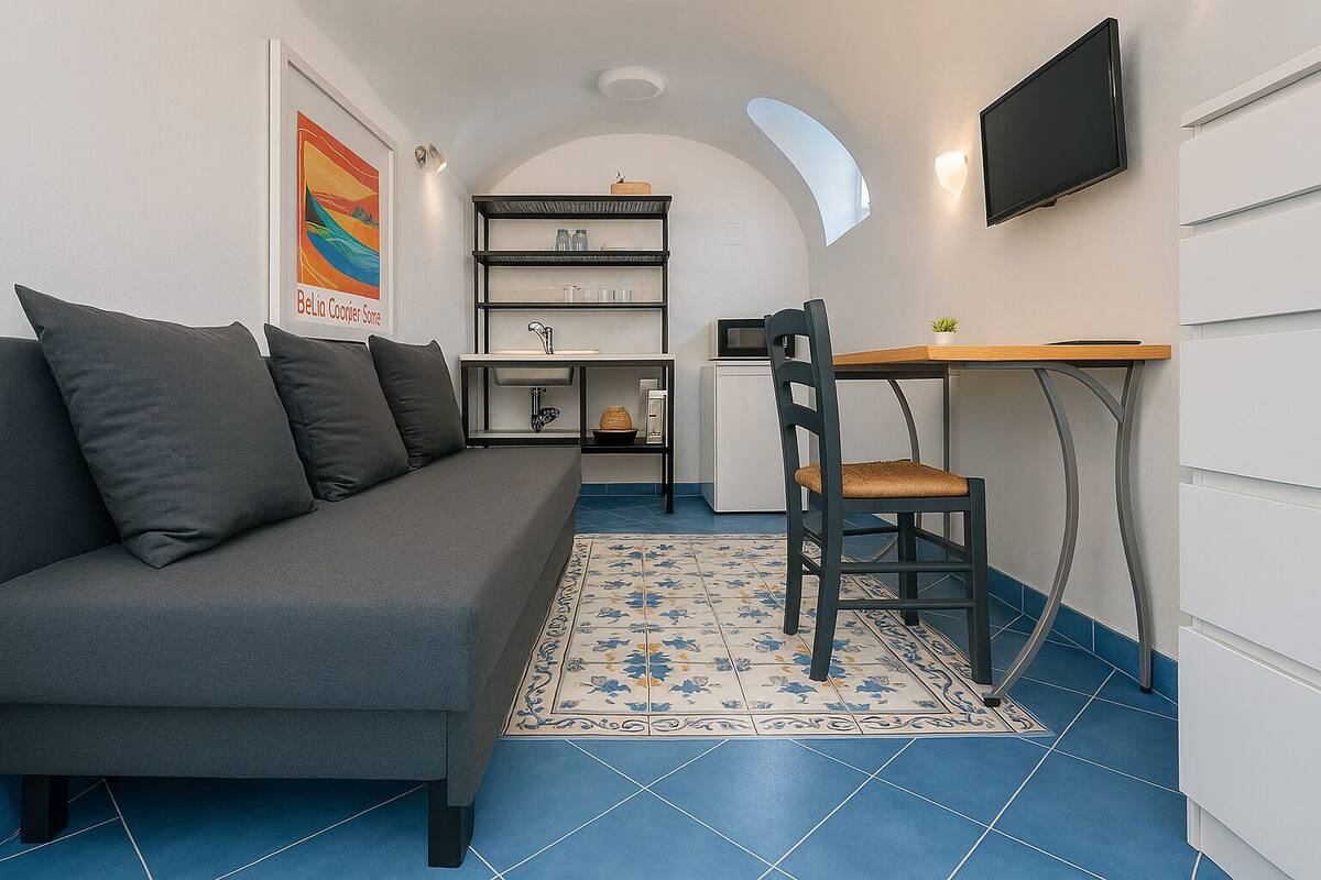 Un espacio habitable compacto con un sofá gris, una mesa de comedor de madera, una silla, un televisor montado en la pared y una zona de cocina con estantes y un refrigerador.