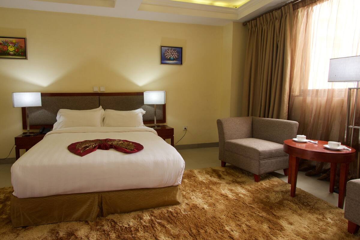 A hotel room with a large bed, two bedside lamps, a chair, a small table, and two paintings on the walls.