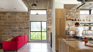 A modern kitchen with a stone wall, wooden cabinets, and a red sofa in a living area. - Pnai - Villa in the Galilee (Rosh Pinna)