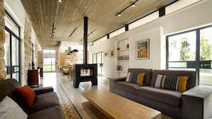 A spacious living room with a large sofa, a wooden coffee table, and a fireplace. There is a kitchen area in the background with a large window. - Pnai - Villa in the Galilee (Rosh Pinna)