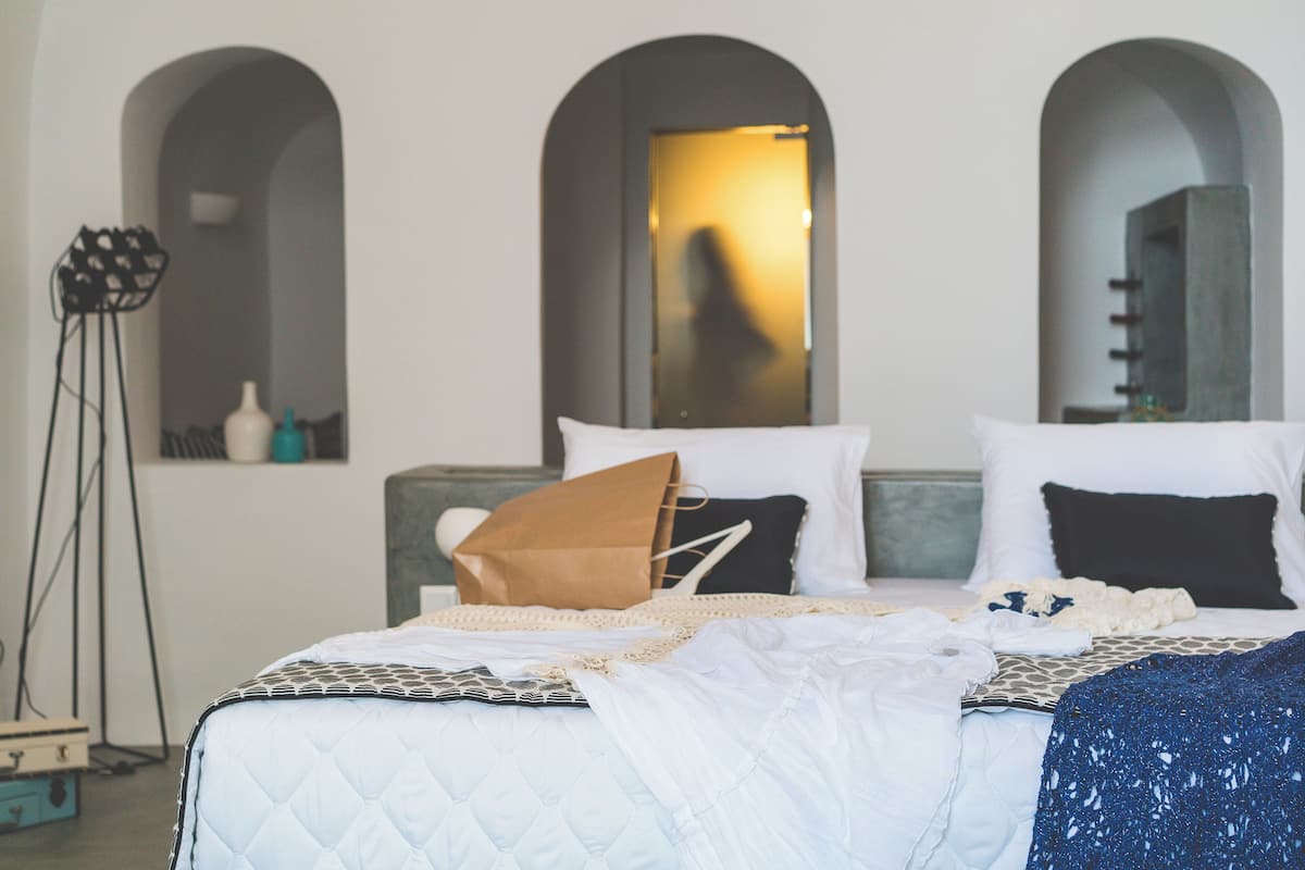 Una cama bien hecha con colcha, almohadas y una bolsa de papel marrón encima. Se observan tres ventanas de arco en la pared detrás de la cama.