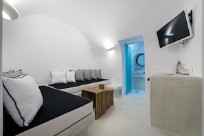 A modern, minimalist bedroom with a curved ceiling, a bed with black and white bedding, a small wooden stool, a white vanity with a mirror, and a mounted TV. - Modernity Suites (Santorini)