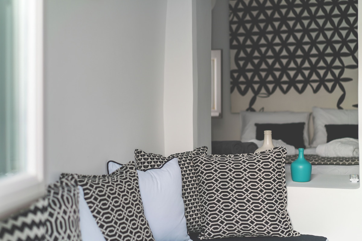 A hotel room with a bed, patterned pillows, a vase, and a geometric wall decoration.