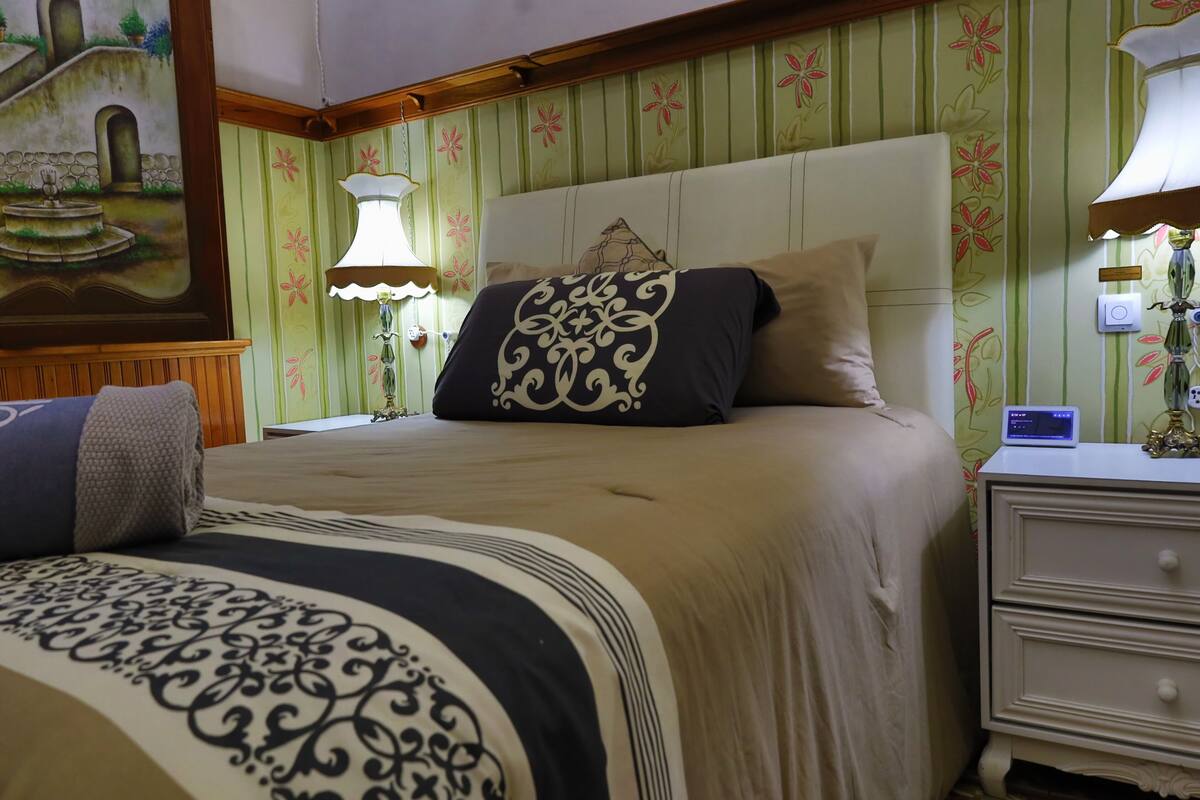 A neatly made bed with a patterned bedspread and pillows, a nightstand with a lamp, and a wall with floral wallpaper.