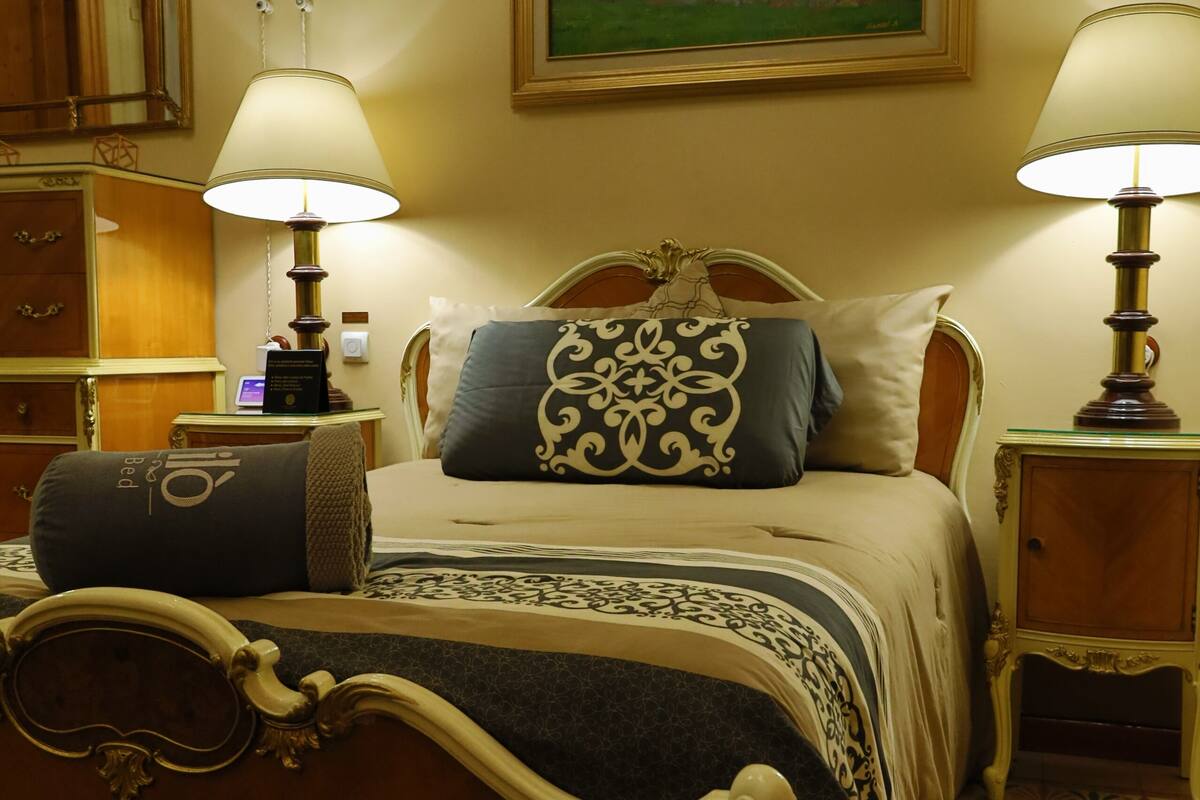 A classic bed with ornate headboard, two bedside lamps, a painting above the bed, and a patterned carpet.