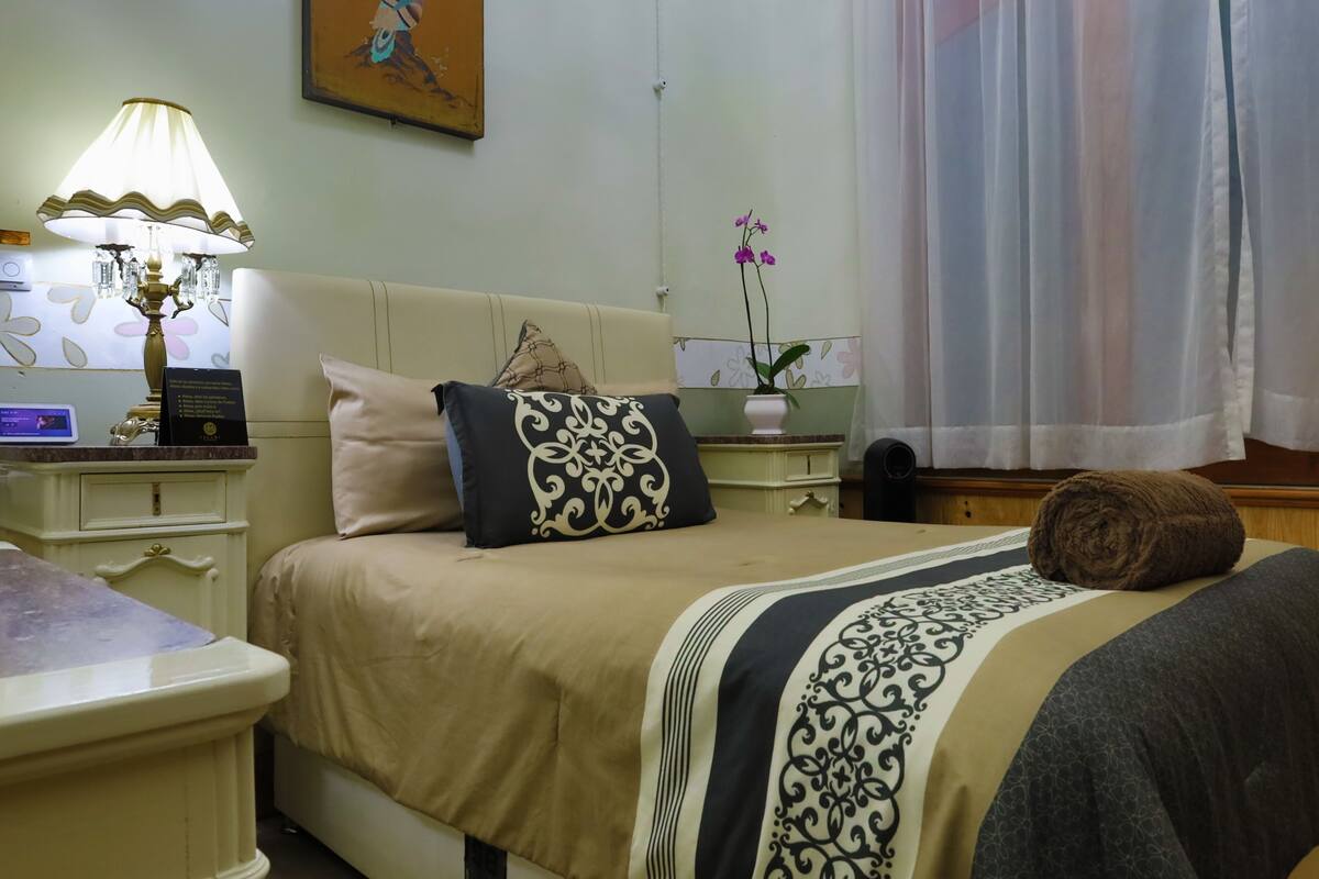 A hotel room with a bed, a nightstand with a lamp, a vase with flowers, and a painting on the wall.