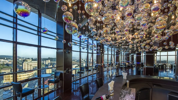 A high-rise bar with a view of the city, large windows, and a ceiling filled with hanging glass spheres.