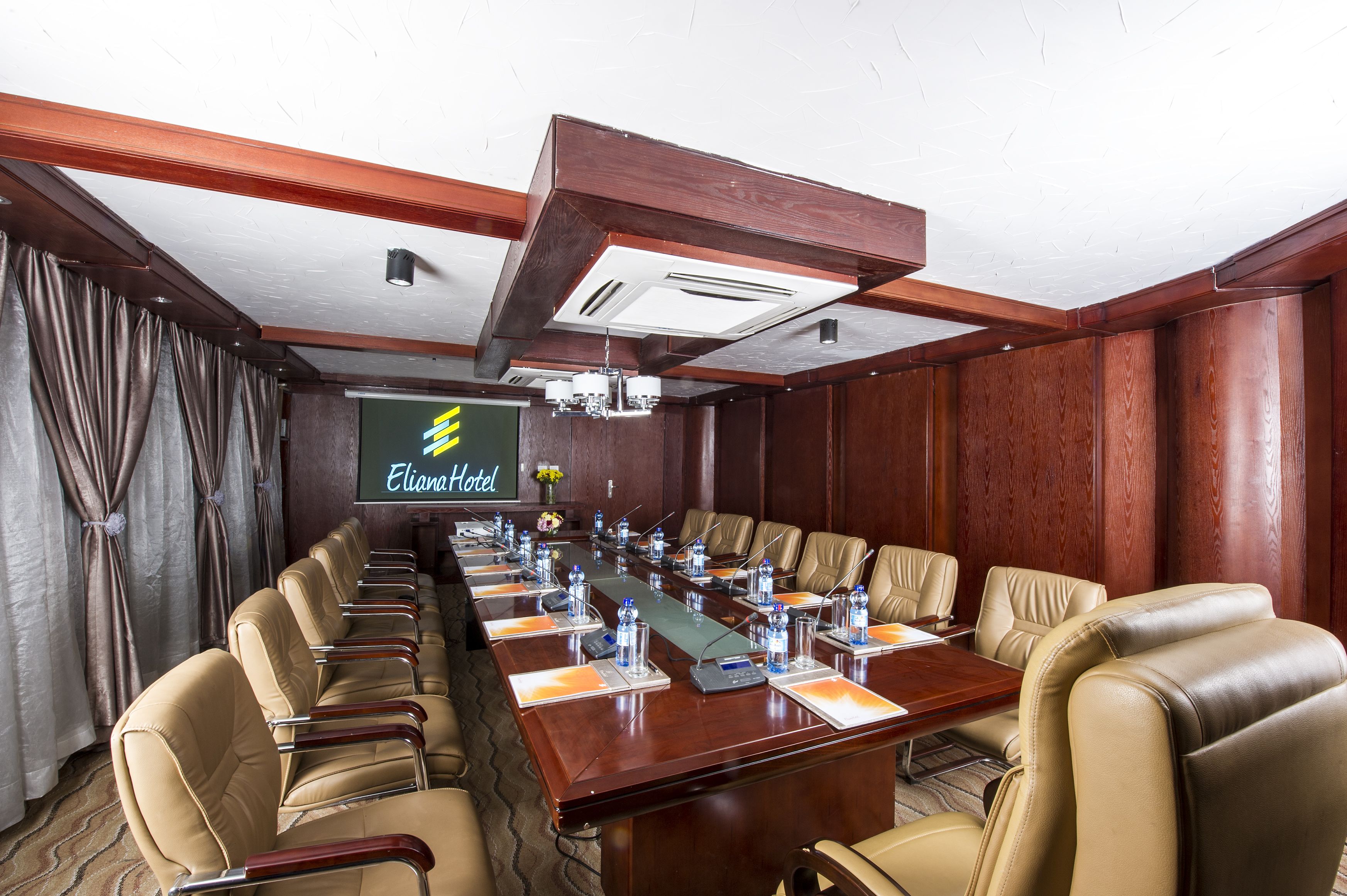 Un salón de conferencias con una larga mesa, sillas de cuero, botellas de agua y una pantalla proyectando "Eliana Hotel".