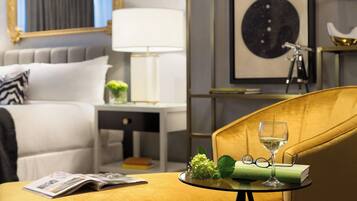 Une chambre d’hôtel avec un lit, une table de chevet avec une lampe, un tableau encadré au mur et une petite table avec un verre de vin, un livre et des lunettes.