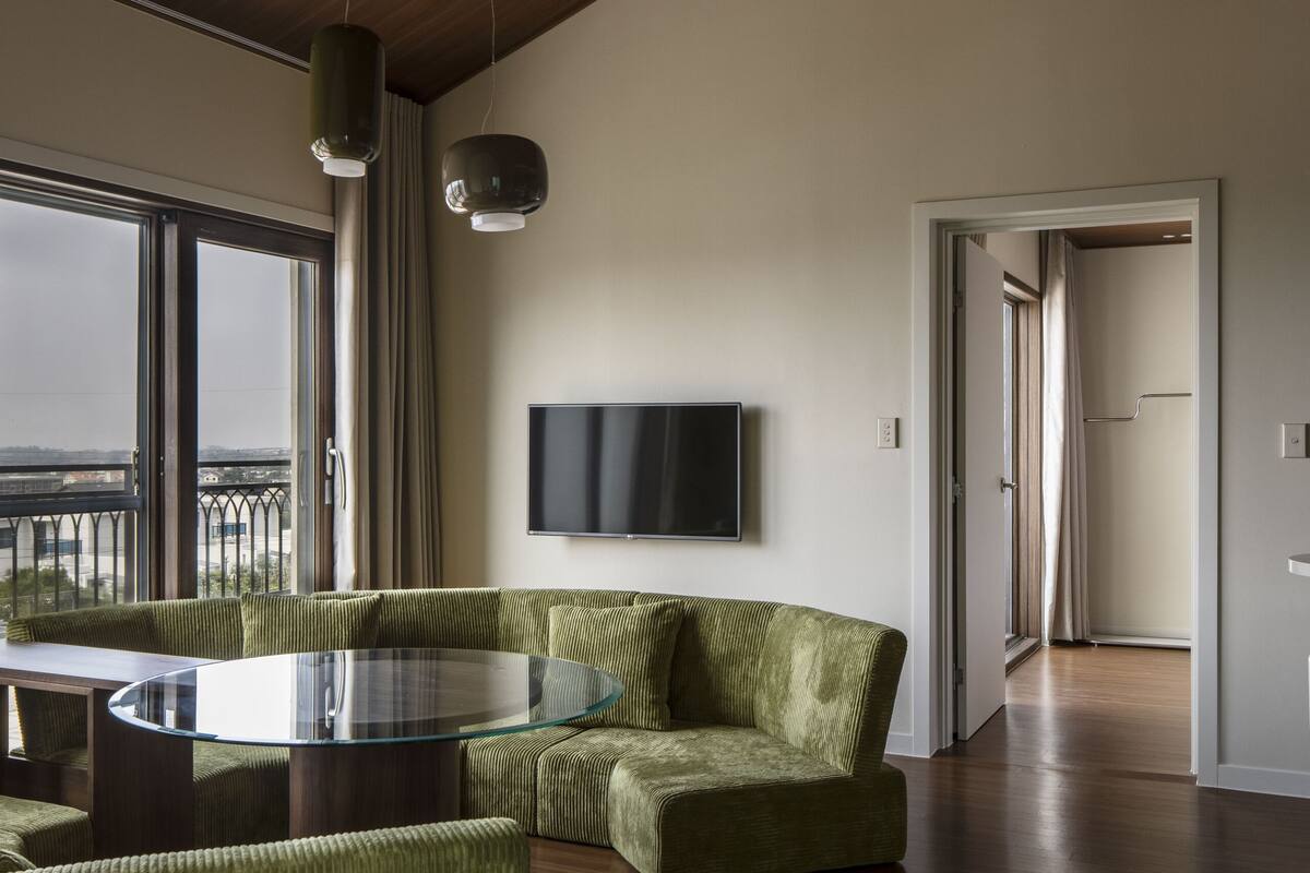 A spacious living area with a large glass table, green upholstered seating, a flat-screen TV mounted on the wall, and a view through large windows.