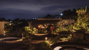 A well-lit outdoor area with a circular water feature, surrounded by rocks and plants, and a building with a tiled roof in the background. - VILLASAGYE (Seogwipo)