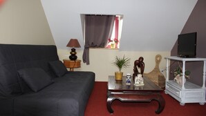 Un salon avec un canapé de couleur sombre, une table basse en bois, un téléviseur posé sur un meuble et une petite fenêtre avec des rideaux.