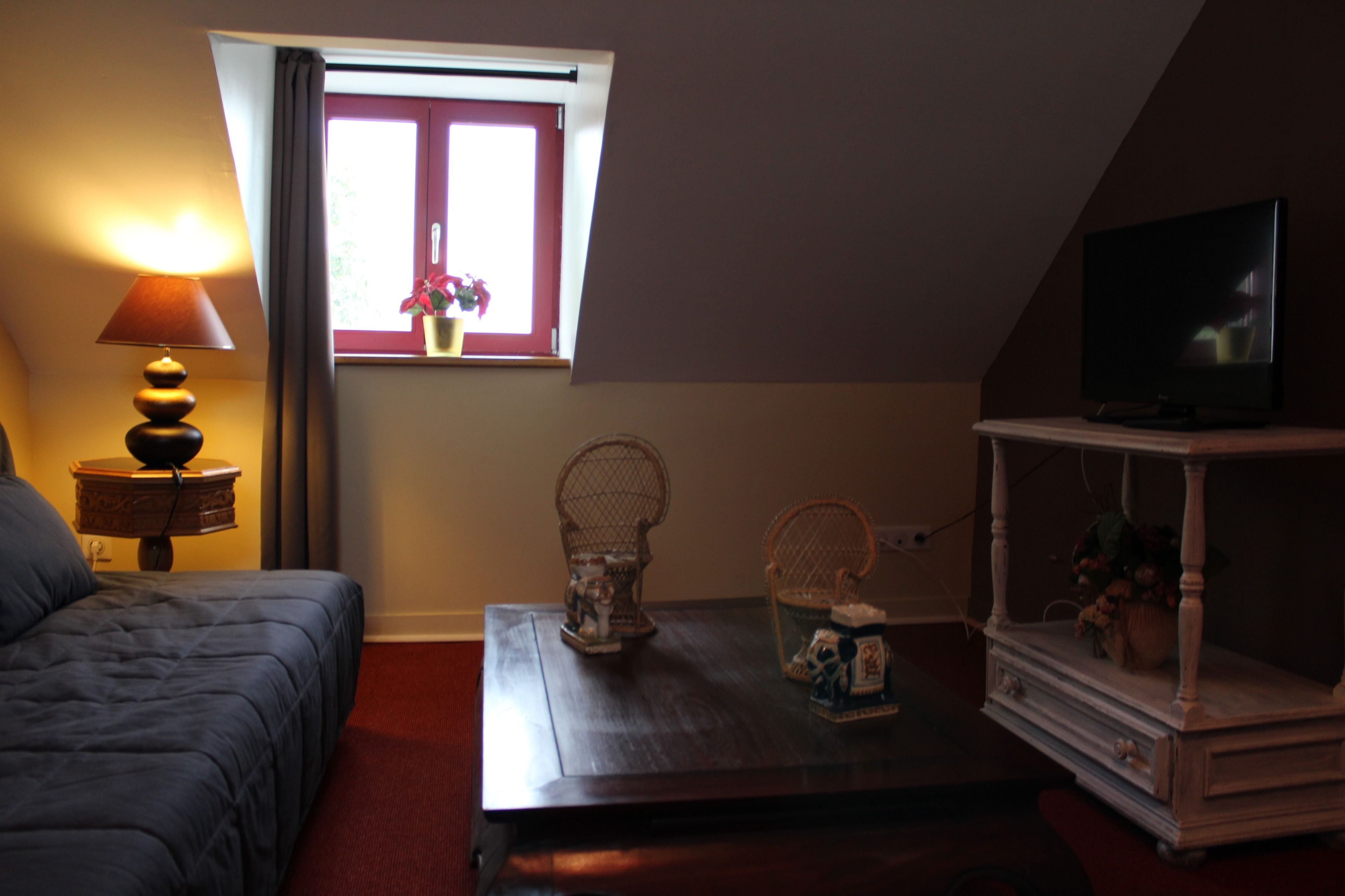 Una habitación con cama, una mesa con objetos decorativos, un televisor sobre un mueble bajo y una ventana con vista a follaje.