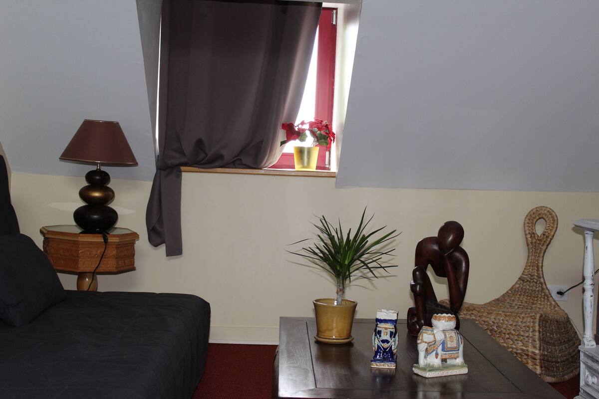 A living room with a sofa, a table with a lamp, a potted plant, a decorative sculpture, and a window with curtains.