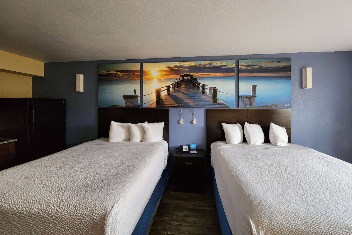 A hotel room with two beds, a painting of a sunset over a pier, and a view of the sea.