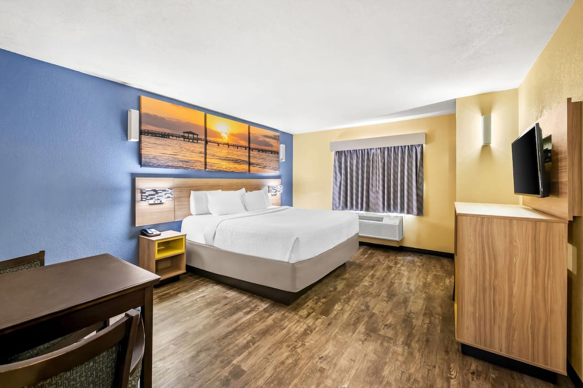 A hotel room with a bed, a desk, a TV, and a painting on the wall.