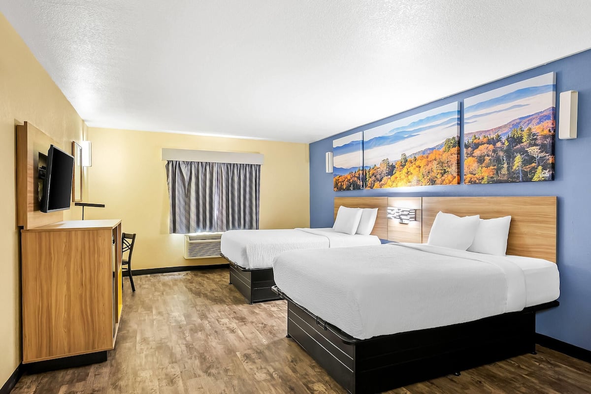 A hotel room with two beds, a desk, a TV, and a large window with curtains.