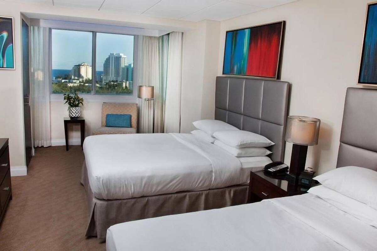A hotel room with two beds, a TV, a desk, and a view of the city.