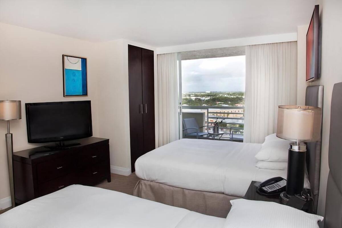 A hotel room with two beds, a TV, a desk with a lamp, a phone, and a balcony with a view of the city.