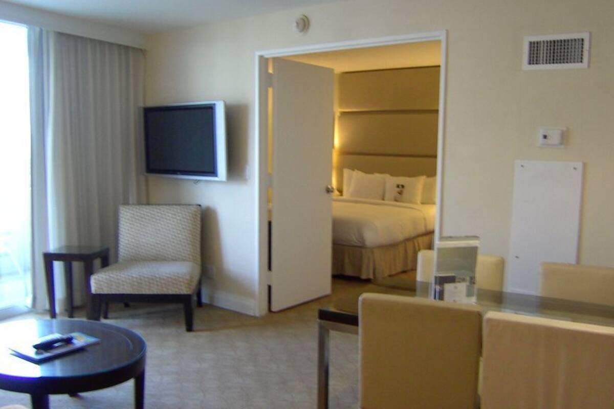 A hotel room with a bed, a TV mounted on the wall, a chair, a small table, and a glass-top table with chairs.
