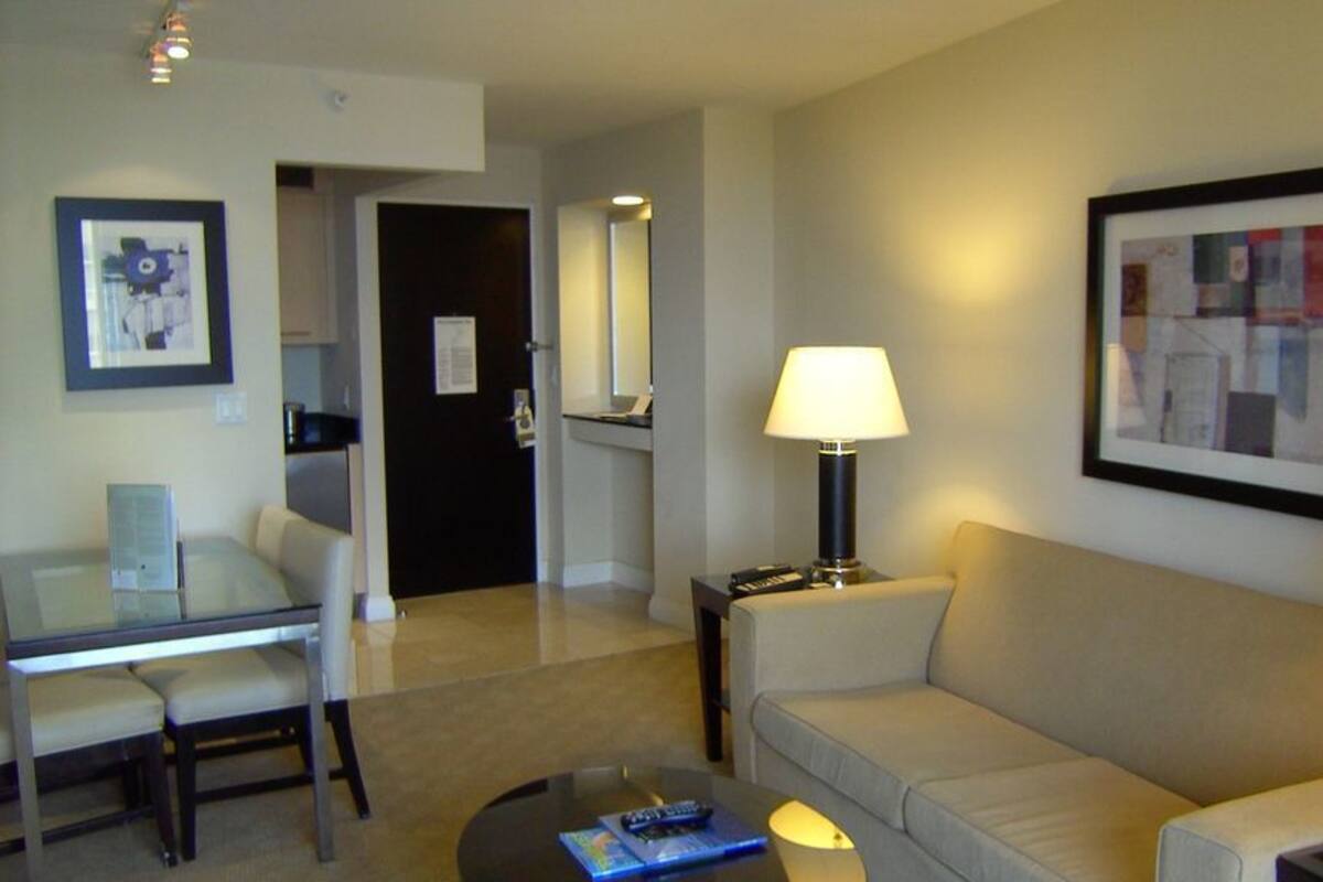 A hotel room with a sofa, a glass table, a coffee table, a lamp, and a framed picture on the wall.
