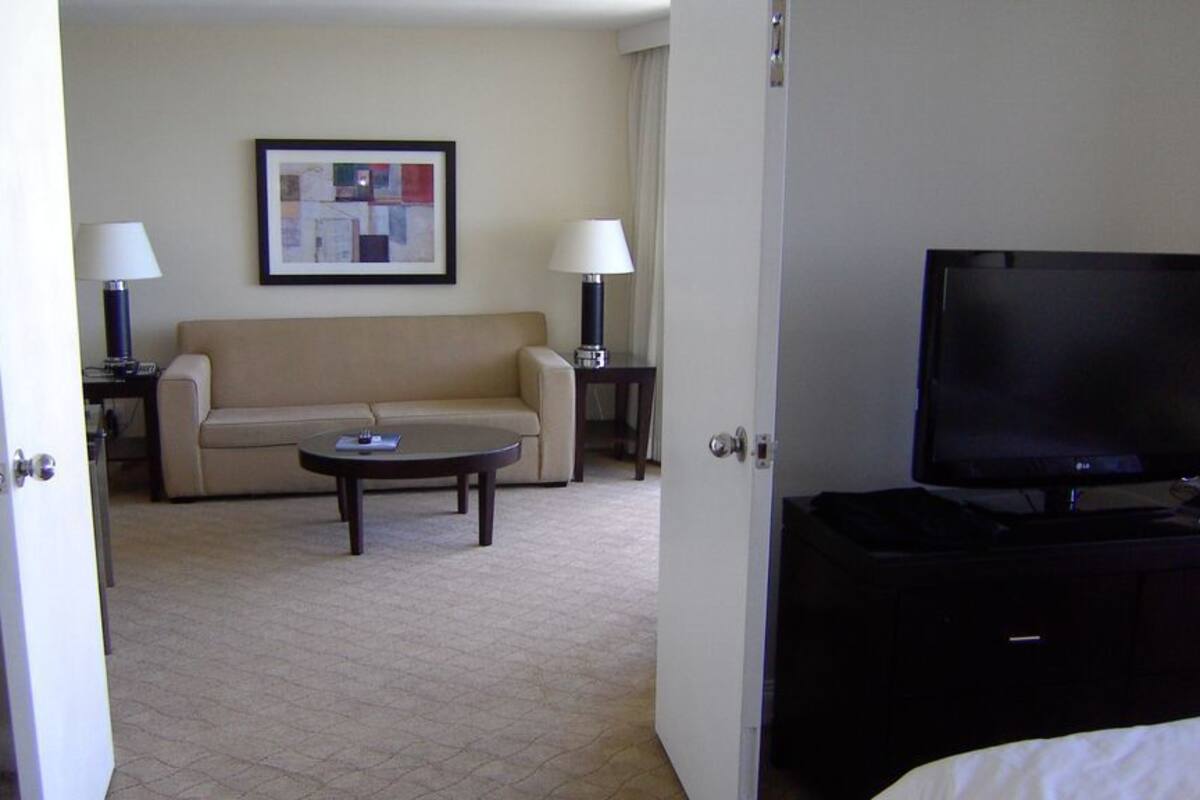 A hotel room with a beige sofa, a coffee table, a flat-screen TV, and a lamp on a side table.