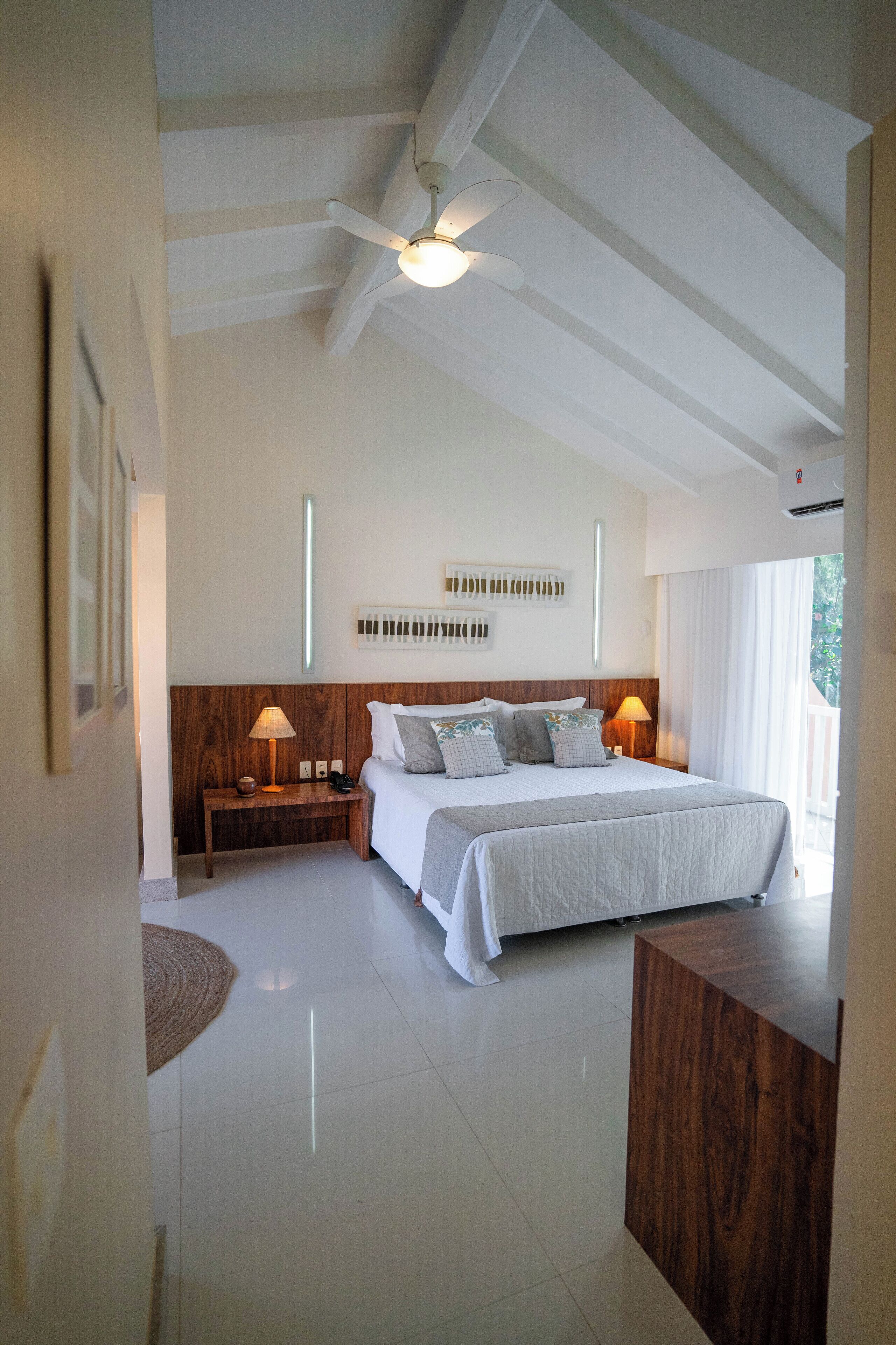 A bedroom with a large bed, two bedside tables with lamps, a ceiling fan, and a view of the outdoors.
