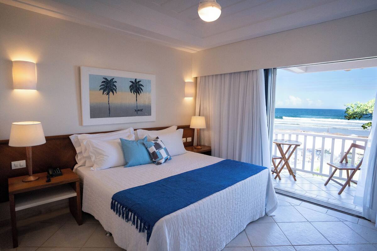 Une chambre d’hôtel avec un grand lit, deux tables de chevet, un tableau représentant des palmiers et un balcon avec vue sur l’océan.