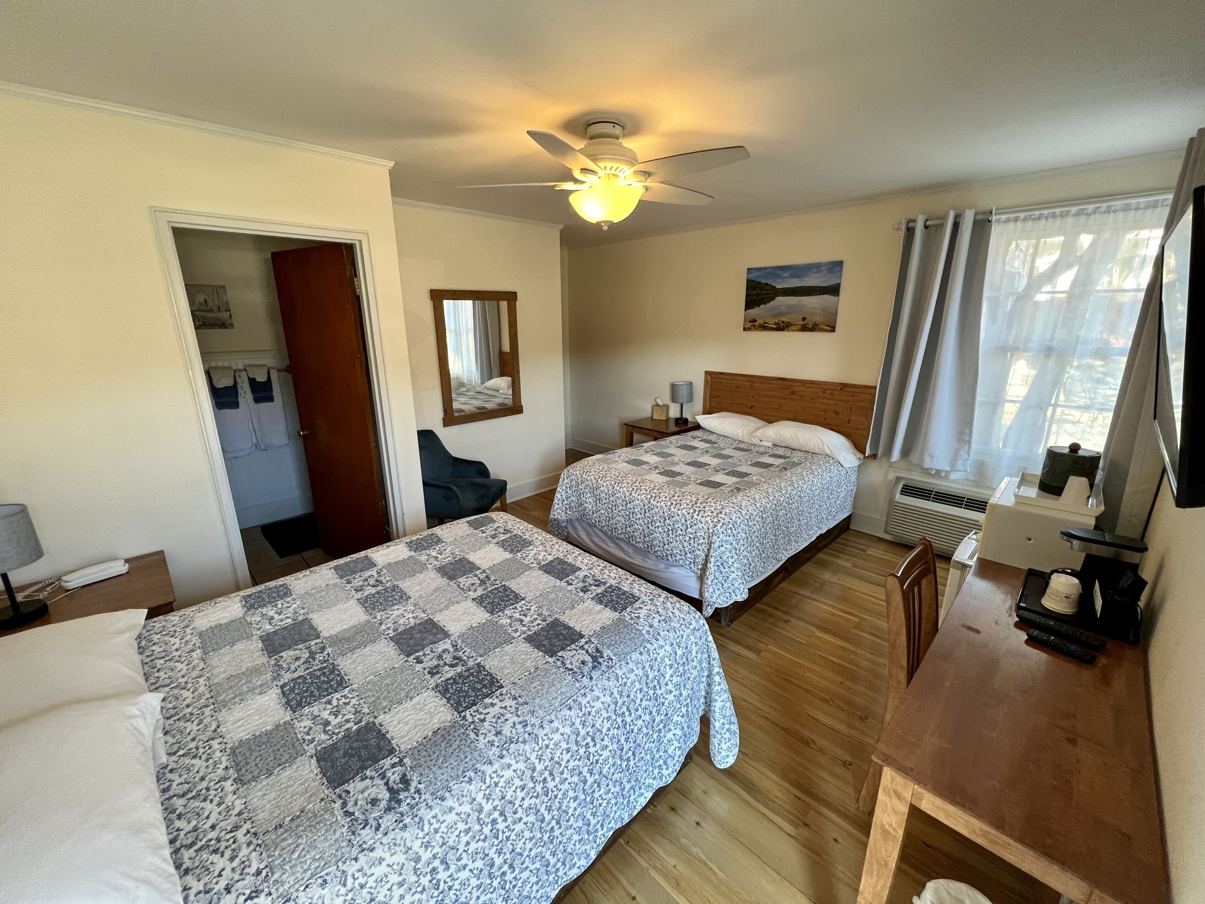 A hotel room with two beds, a ceiling fan, a painting, a window with curtains, and a desk with a lamp.