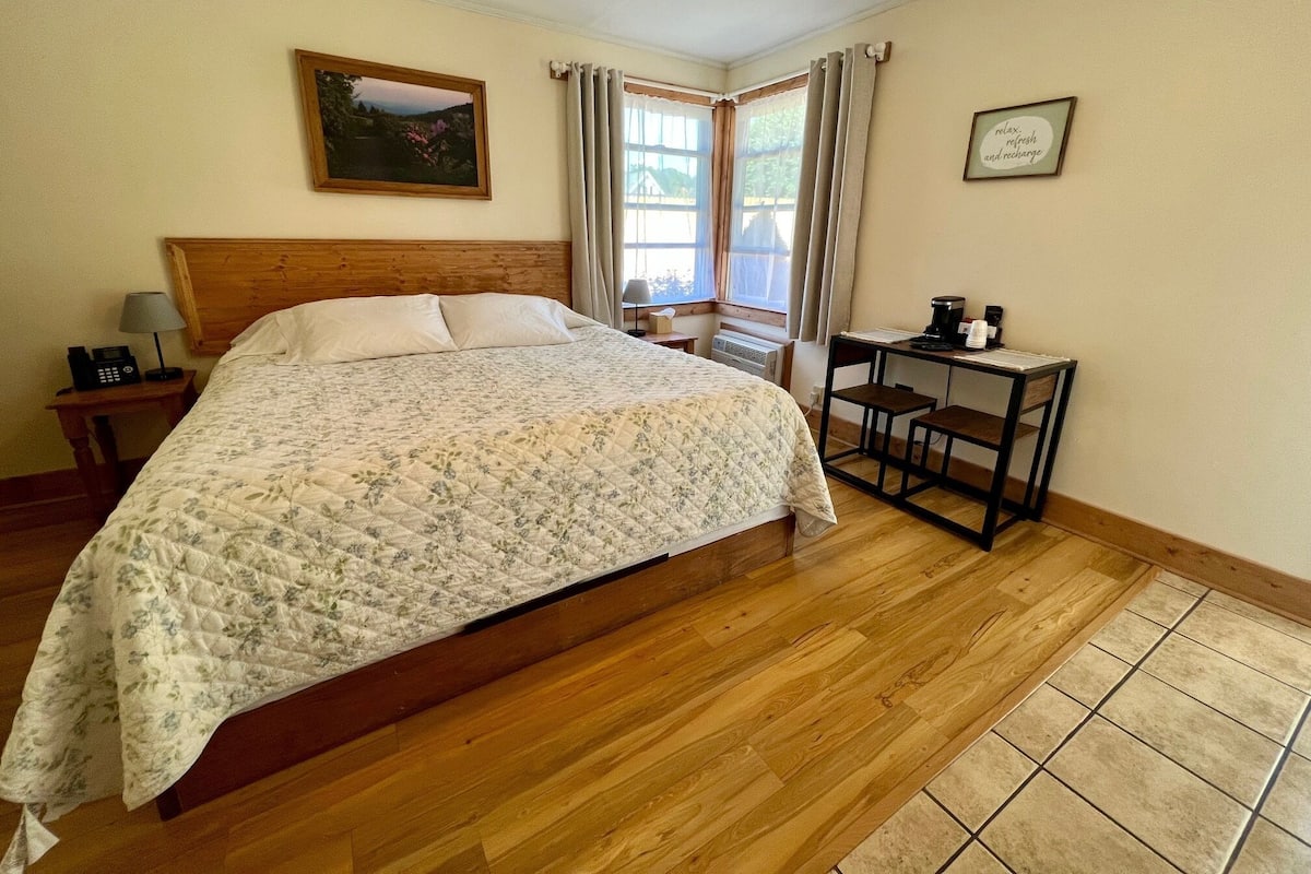 A bedroom with a wooden bed, a desk with a chair, a telephone, and a picture frame on the wall.