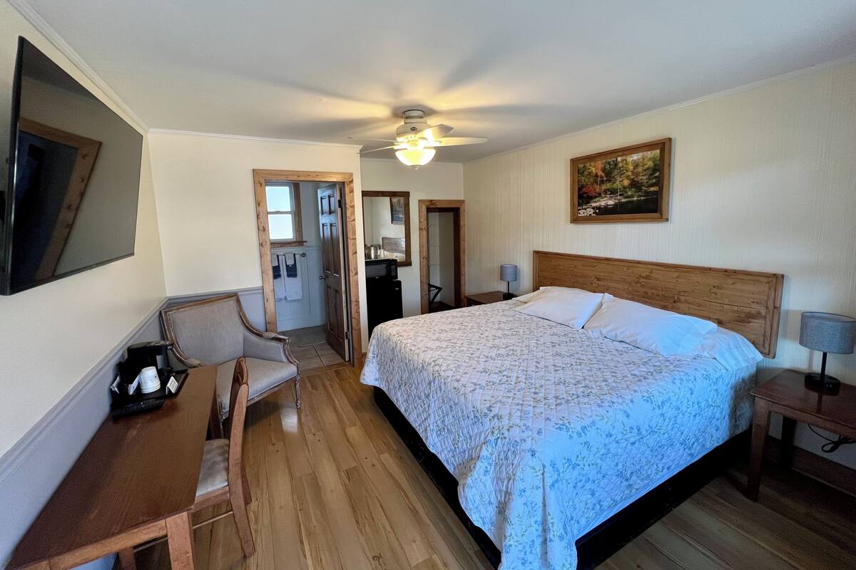 A bedroom with a bed, a desk, a chair, a ceiling fan, and a painting on the wall.