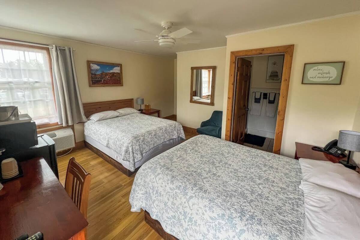 A hotel room with two beds, a desk, a chair, a ceiling fan, and a door leading to a bathroom.
