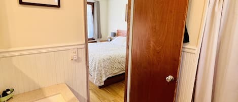 A bathroom with a sink, mirror, and shower area. A door leads to a bedroom with a bed and a ceiling fan.