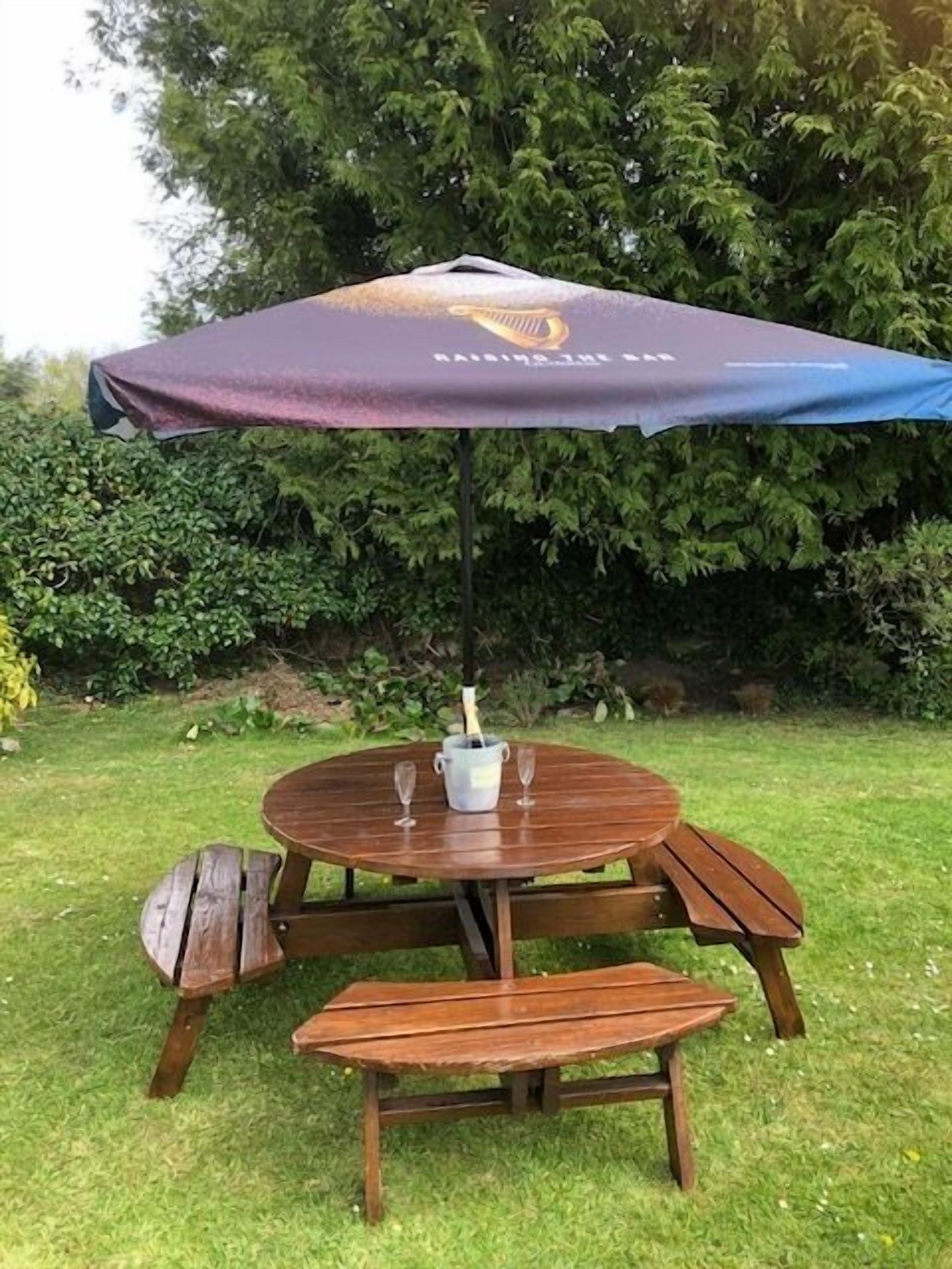 A wooden outdoor table with a bench, a bottle of champagne, and two glasses under a purple umbrella.