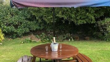 A wooden outdoor table with a bench, a bottle of champagne, and two glasses under a purple umbrella.