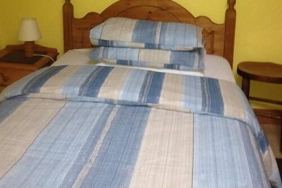 A bed with a striped bedspread and pillows, a wooden headboard, a nightstand with a lamp, and a small wooden stool.