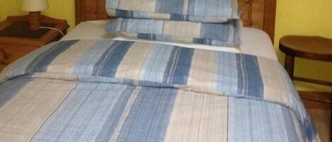 Una cama con colcha a rayas y almohadas, cabecera de madera, mesita de noche con lámpara y un taburete pequeño de madera.
