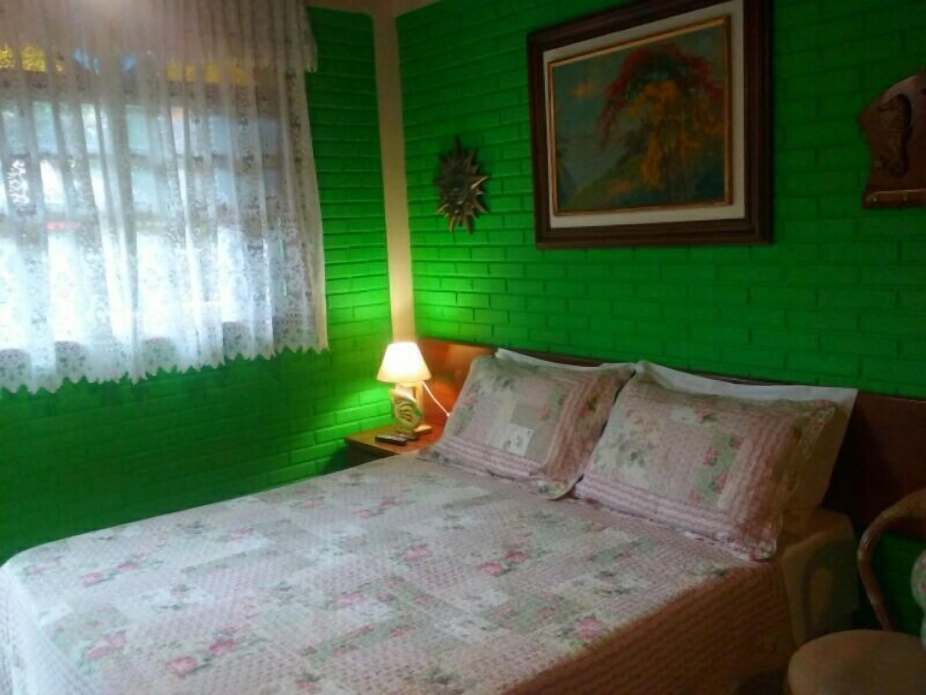 Una habitación con pared de ladrillo verde, una cama con ropa de cama floral, una mesita de noche con una lámpara y un cuadro en la pared.