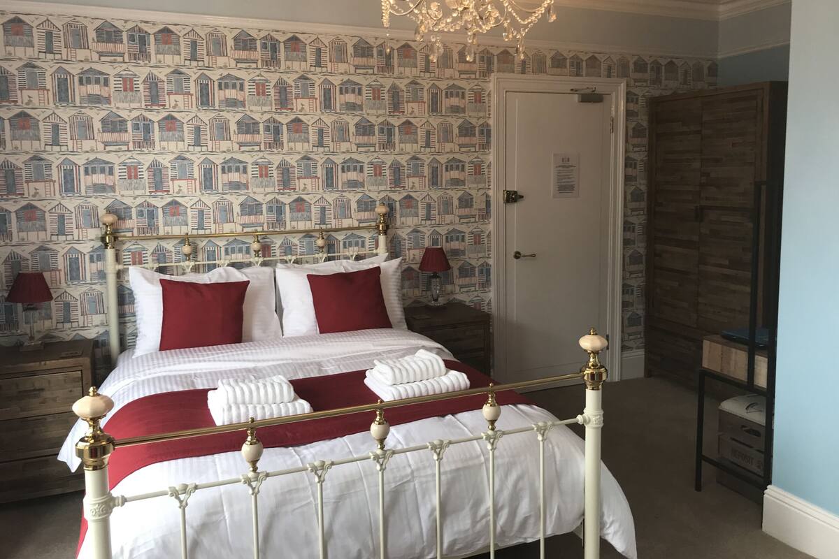 Cama bem arrumada com roupa de cama branca e vermelha, um candelabro acima e papel de parede estampado com desenhos de casas.