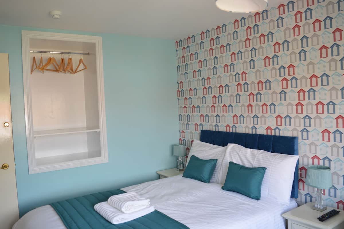 Quarto com uma cama, mesa de cabeceira, luminária, janela com cortineira e parede com padrão de casa.